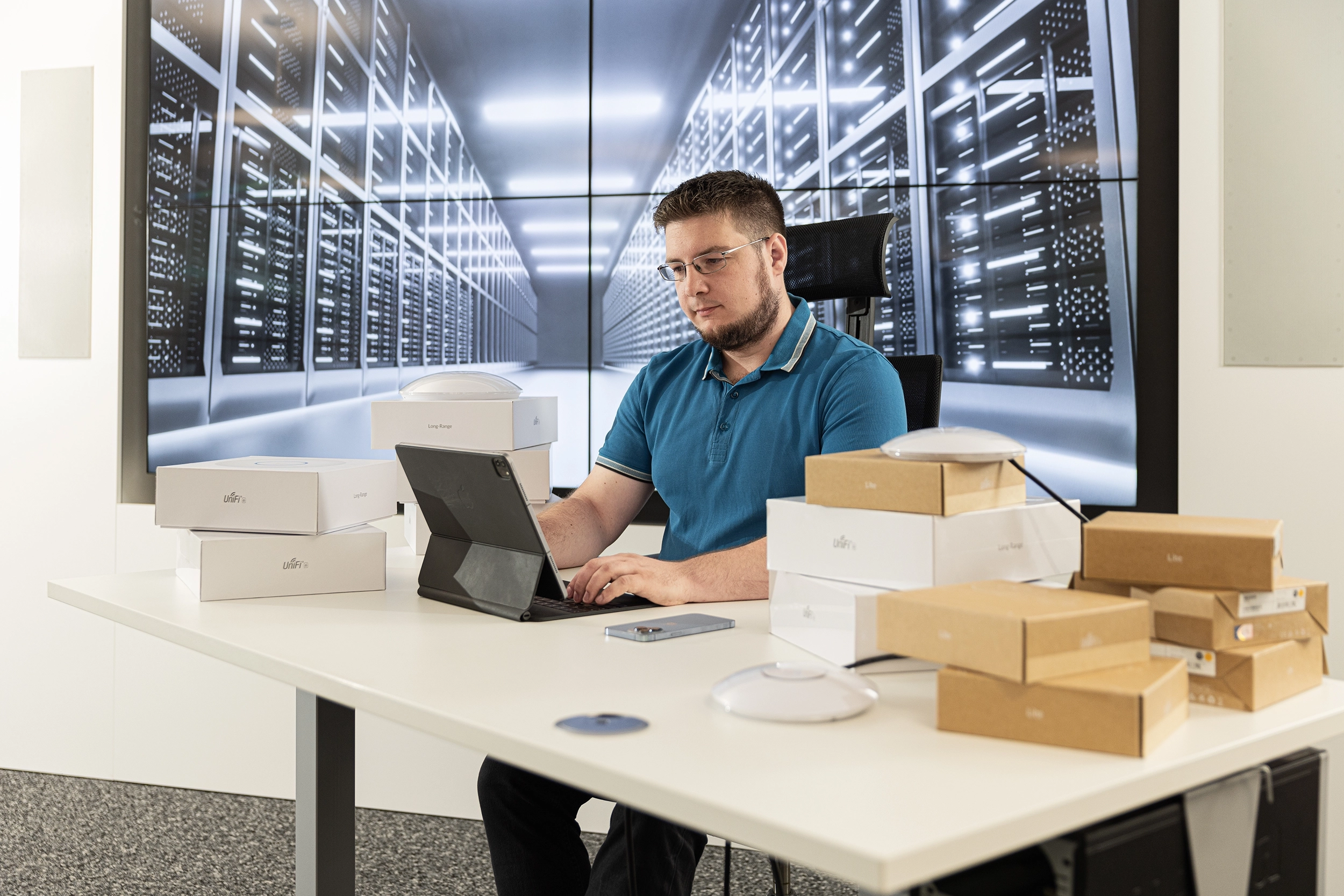03_IMG_0737 – IT Lösungen Mann an Schreibtisch arbeitet am Laptop, umgeben von Routerboxen, mit großem Bildschirm im Hintergrund, der Serverraum zeigt.