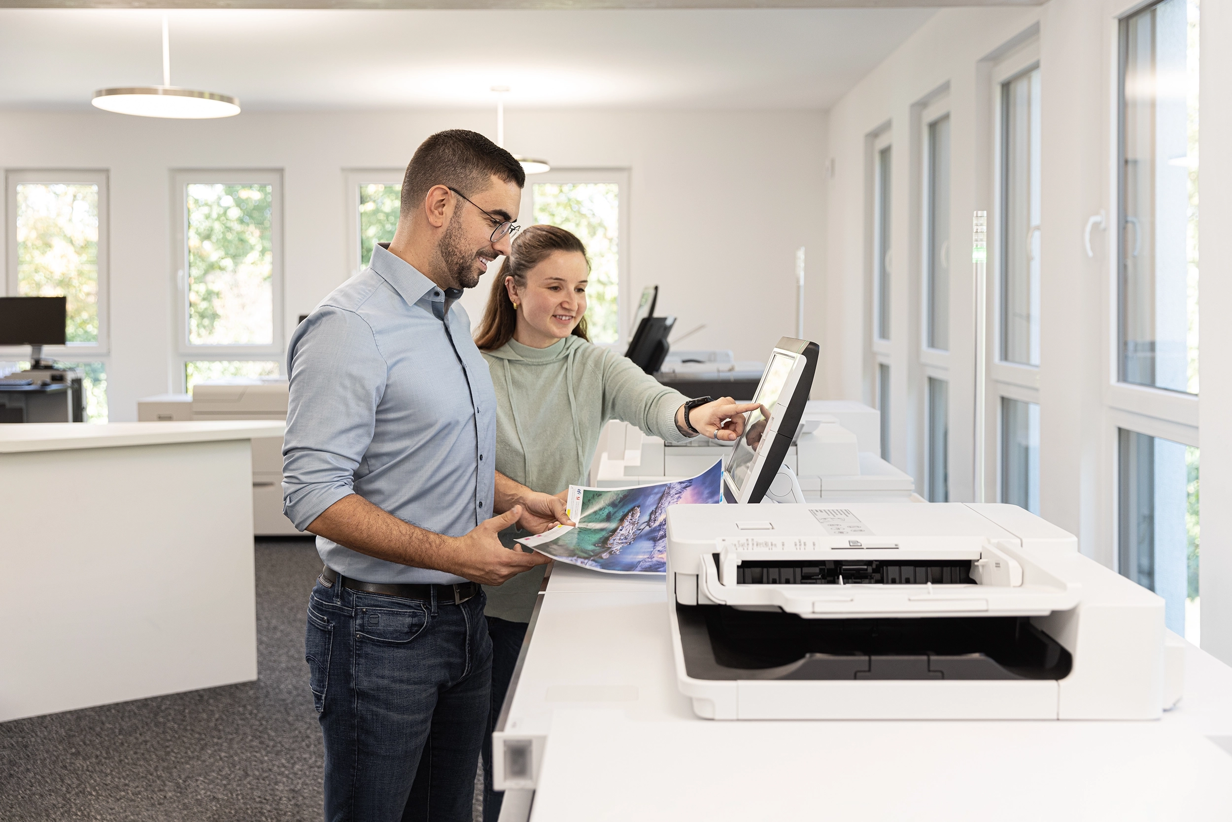 06_IMG_1510 – Produktionsdruck Zwei Personen nutzen in einem Büro Druckernetzwerk. Eine zeigt auf das Touchscreen-Display, während die andere ein Foto hält.