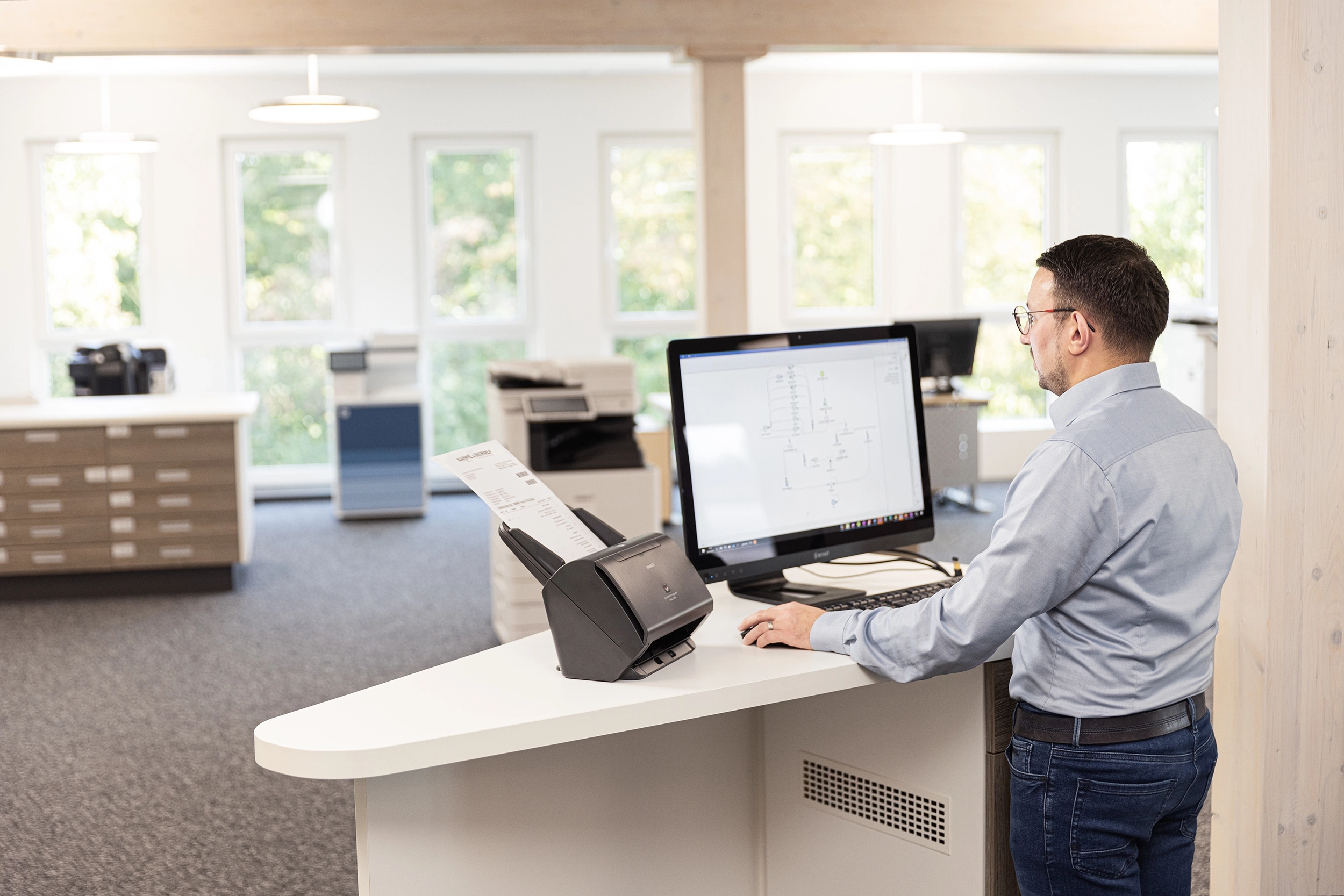 08_IMG_2083 – Dokumentenmangement Mann im Büro arbeitet an Computer mit Scanner am Schreibtisch, helle Fenster im Hintergrund.