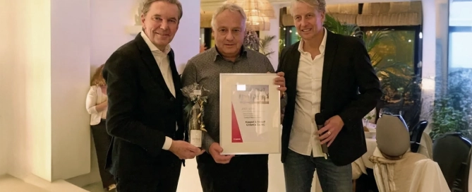 Drei Männer in Anzügen halten eine Urkunde und eine Flasche in einem elegant beleuchteten Raum in die Kamera.