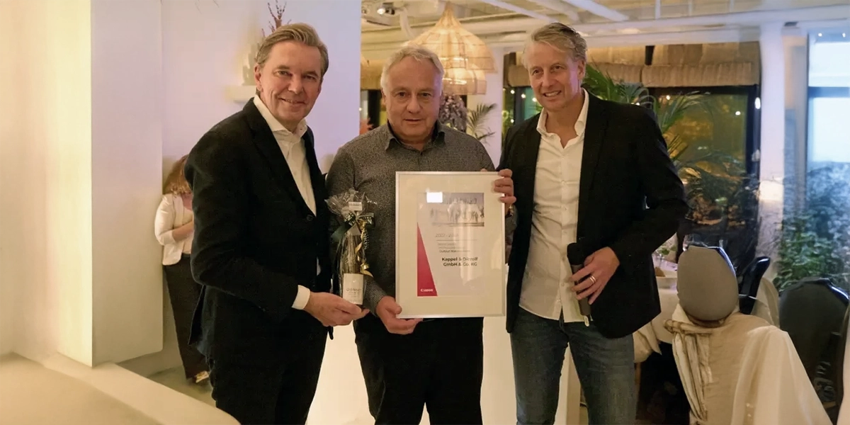 Kappel-Dierolf-Blog_Canon_Partner_Convention_2024 Drei Männer in Anzügen halten eine Urkunde und eine Flasche in einem elegant beleuchteten Raum in die Kamera.