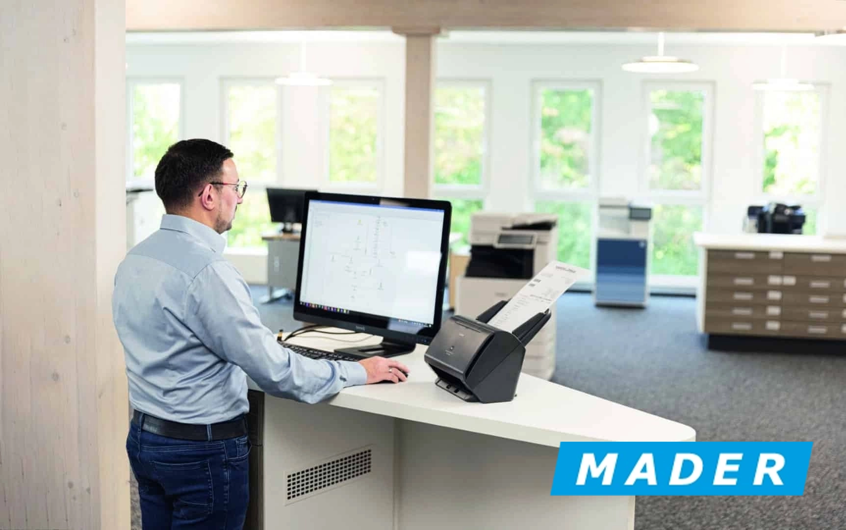 Mitarbeiter steht vor Computer in modernem Büro, nutzt Scanner für Dokumenteneingabe. Firmenlogo im Vordergrund.