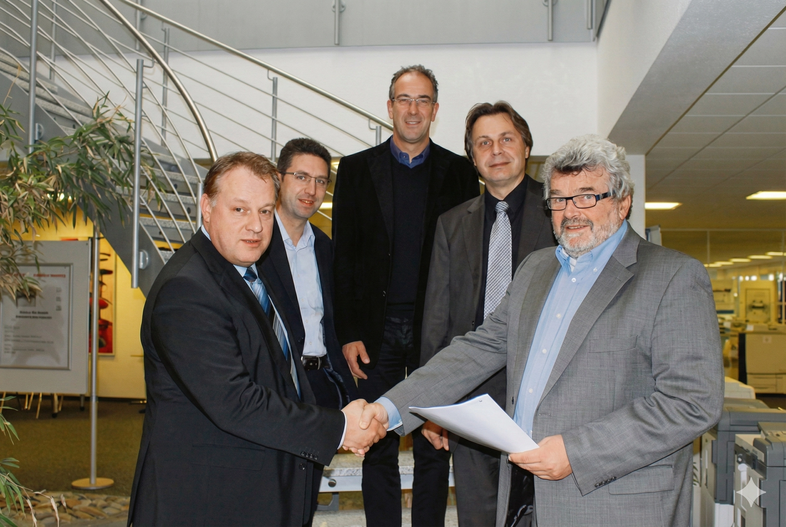 Kappel-und-Dierolf_uebernimmt_Kawa Fünf Männer in Anzügen stehen zusammen. Zwei Männer im Vordergrund schütteln die Hände und halten ein Dokument.