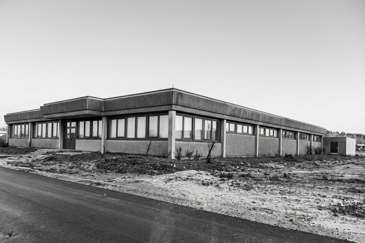 Kappel_1983_BüroNUEZIZIS Graues, rechteckiges Betongebäude mit vielen Fenstern und einer Eingangstür, umgeben von karger Landschaft.