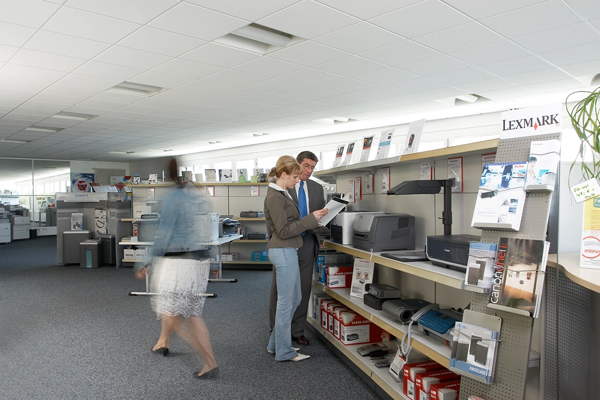 Kappel_Büroeinrichtungshaus Zwei Personen betrachten Druckermodelle in einem Büroausstellungsraum, während eine unscharfe Person im Vordergrund vorbeigeht.