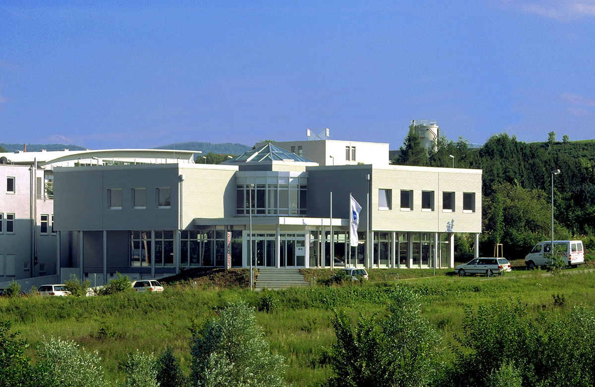 Kappel_Firma_Kirchheim Modernes Bürogebäude mit Glasdach und mehreren Fahrzeugen davor, umgeben von grüner Vegetation und klarem Himmel.