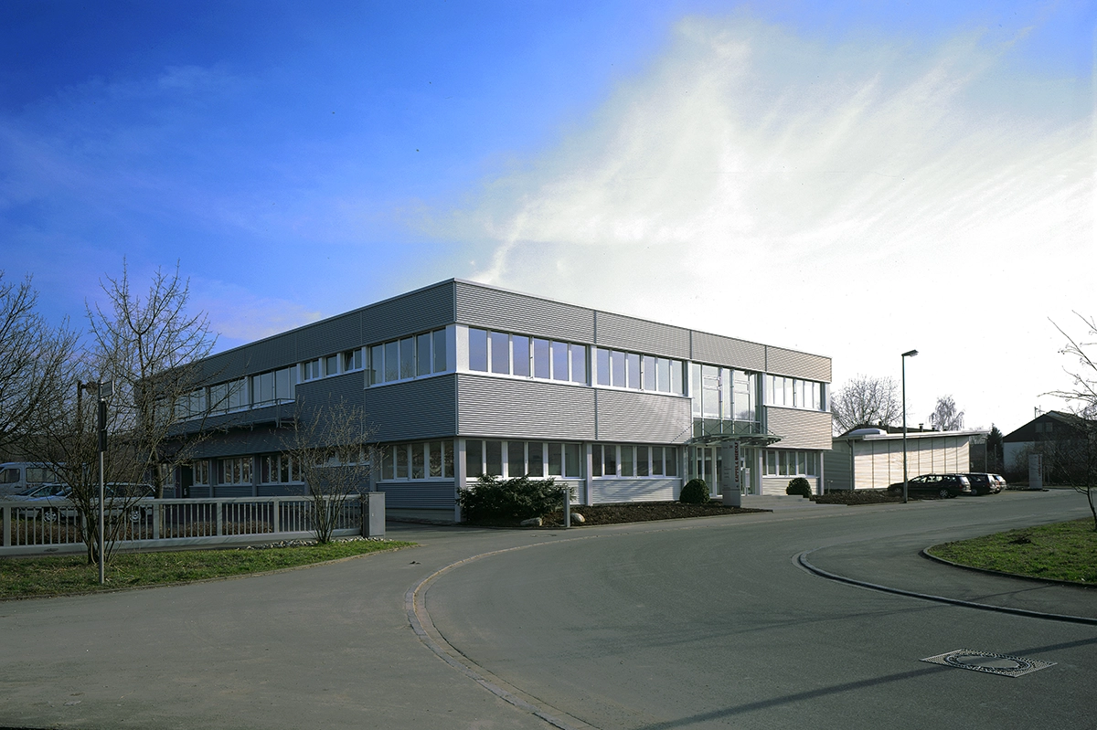 Kappel_Gebäude_Zizishausen Modernes zweistöckiges Bürogebäude mit großen Fenstern, umgeben von einer Straße und kahlen Bäumen unter klarem Himmel.