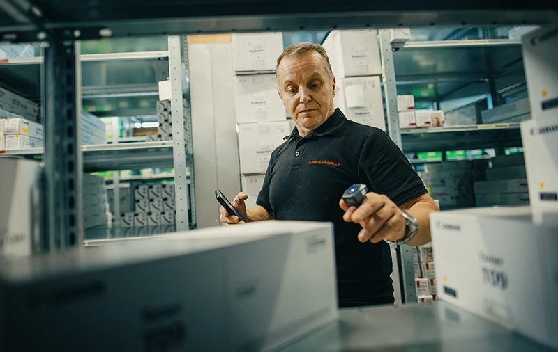 Mann im Lager scannt Kartons mit Handscanner, umgeben von Regalen mit Produkten.