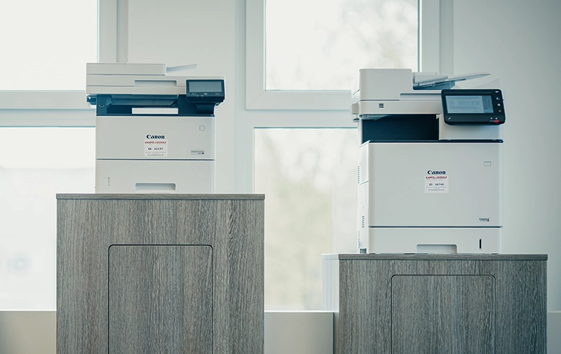 Zwei Kopierer auf grauen Holzschränken vor Fenstern in einem hellen Büro.