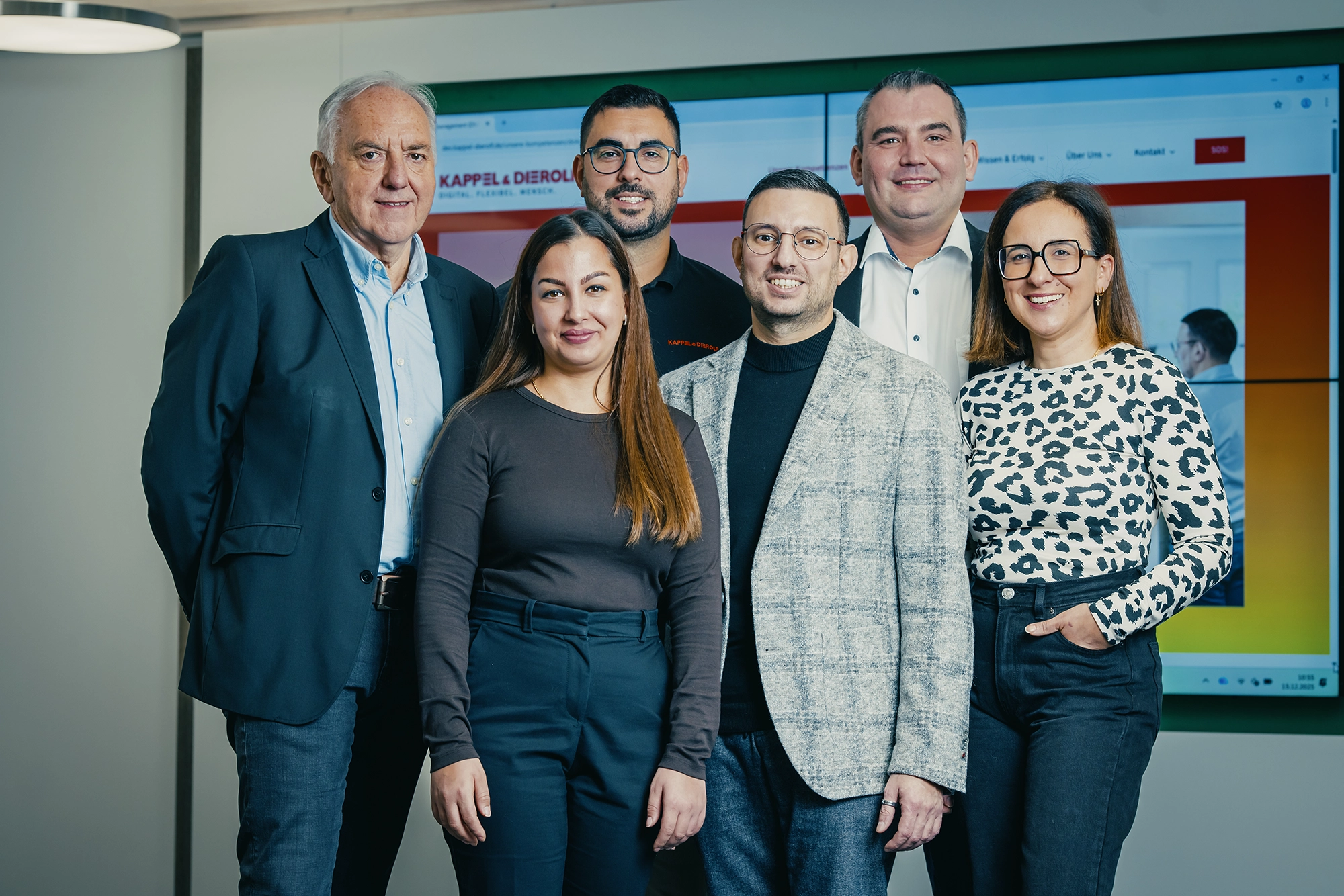 Sechs Personen stehen lächelnd in einem Büro vor einem Monitor, der die Website Kappel & Dierolf zeigt.