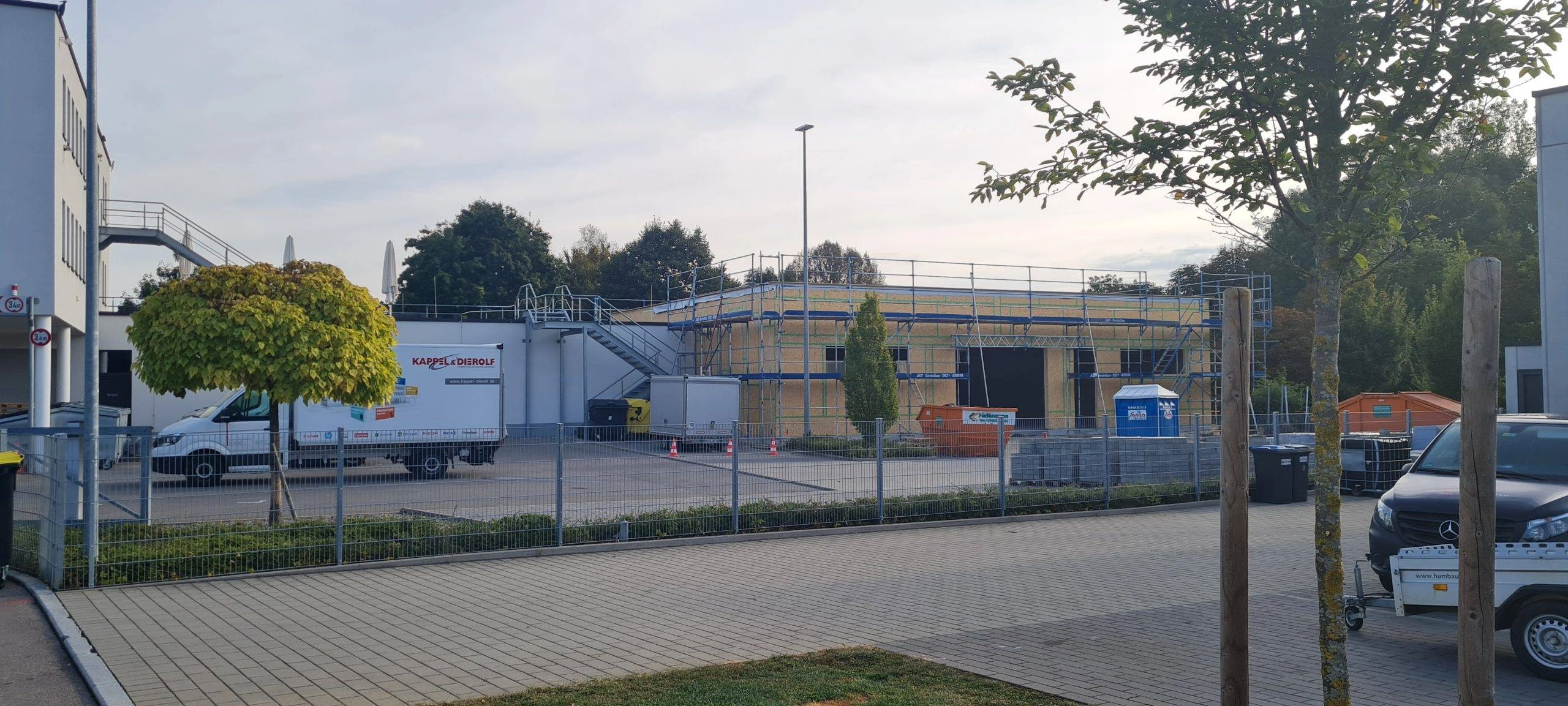 anbau2 Bauplatz mit Gerüst und Lkw vor teilweise errichteter Halle, umgeben von Bäumen und Bauzäunen, bei hellem Tageslicht.