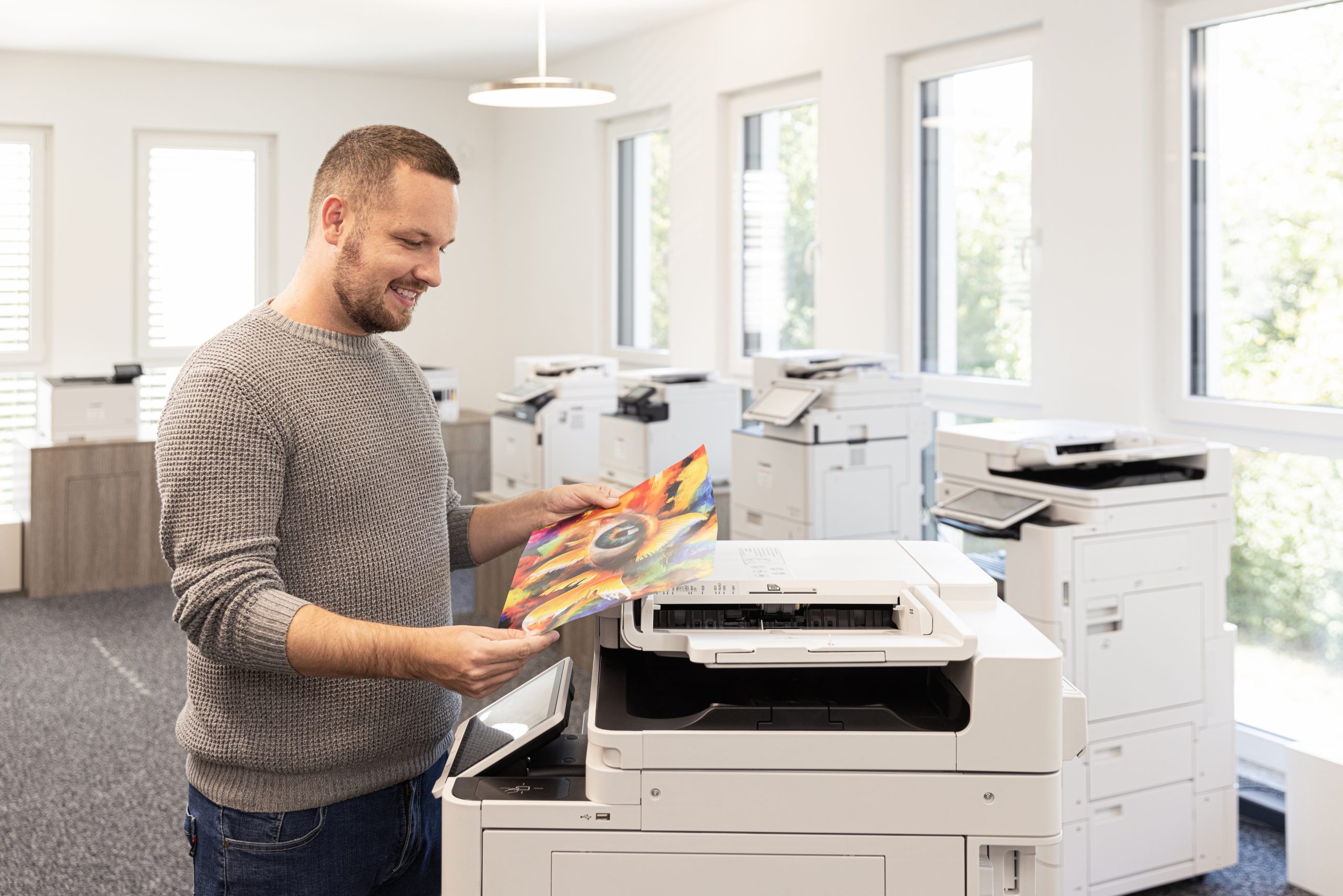 Mann in grauem Pullover betrachtet ein farbenfrohes Druckbild in einem Büro mit mehreren Druckern.