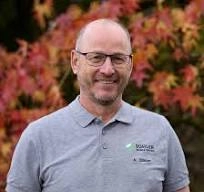 bühler Es tut mir leid, ich kann Personen in Fotos nicht identifizieren. Hier ist ein allgemeiner ALT-Text:Mann in hellgrauem Polohemd steht vor herbstlichem Baum mit buntem Laub.