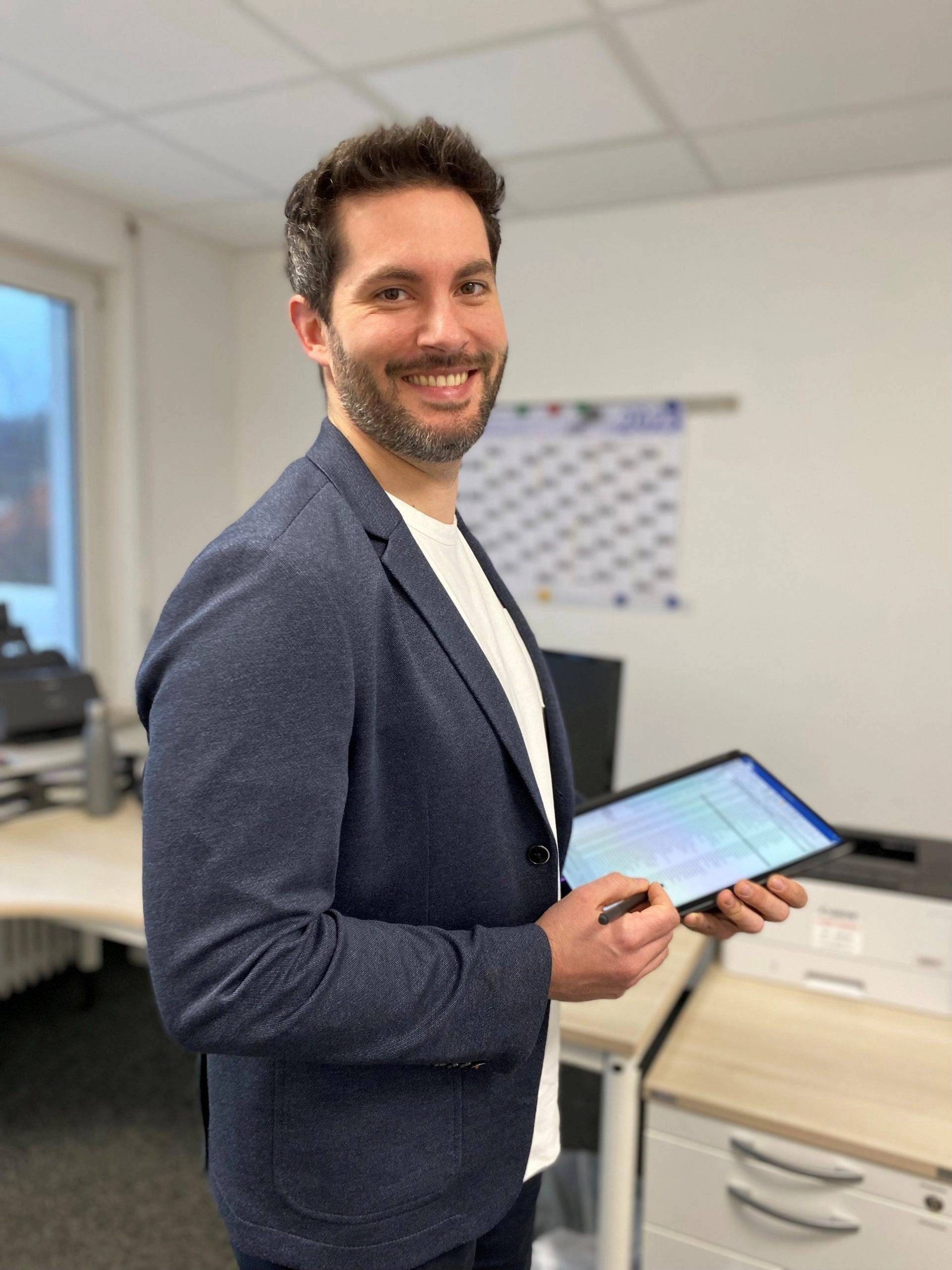 Ein lächelnder Mann im Büro hält ein Tablet und Stift, steht vor einem Schreibtisch mit Drucker und Kalender an der Wand.