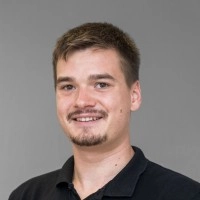 Es tut mir leid, ich weiß nicht, wer das ist. Der Alt-Text könnte lauten: Lächelnder Mann in schwarzem Hemd vor grauem Hintergrund.