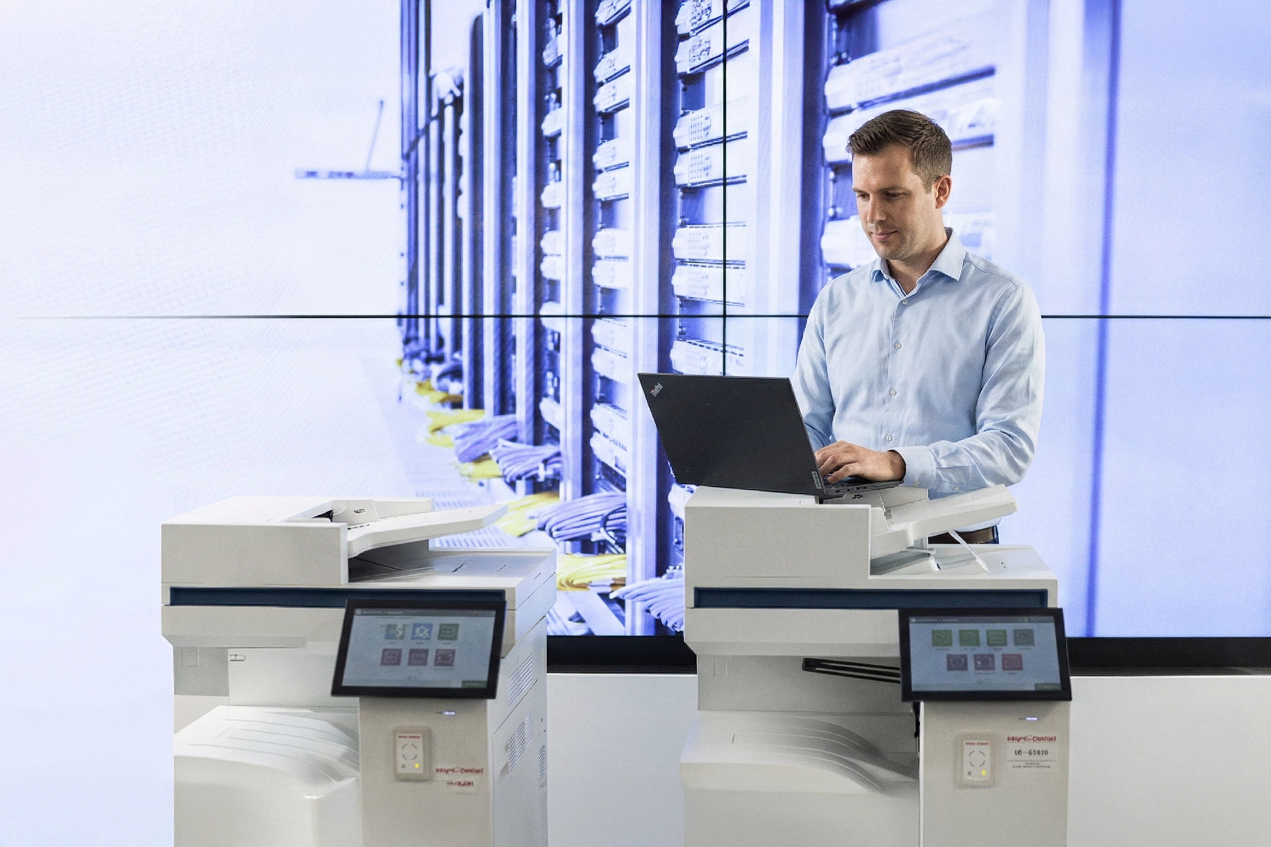 kappel-dierolf-header-sicherheit Mann in blauem Hemd arbeitet mit Laptop auf Drucker vor Serverraum-Hintergrund. Büro-Umgebung, Fokus auf Technik.