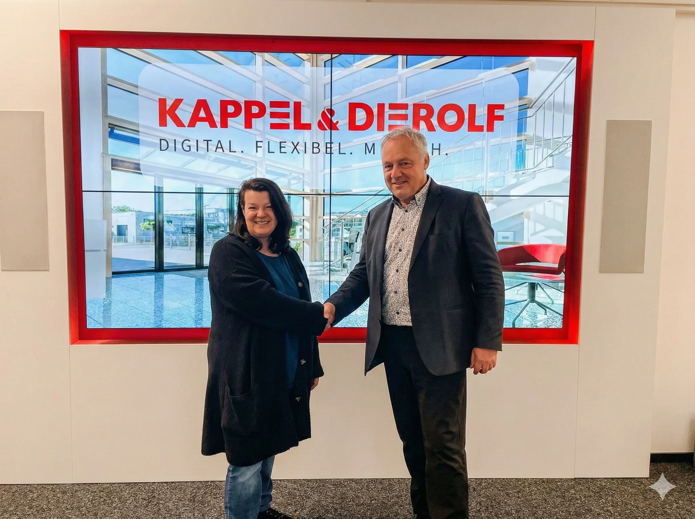 uebernahme_ANKA_Drucker_Service Zwei Personen schütteln Hände vor einem großen Bildschirm mit Kappel & Dierolf Werbung im Büro.
