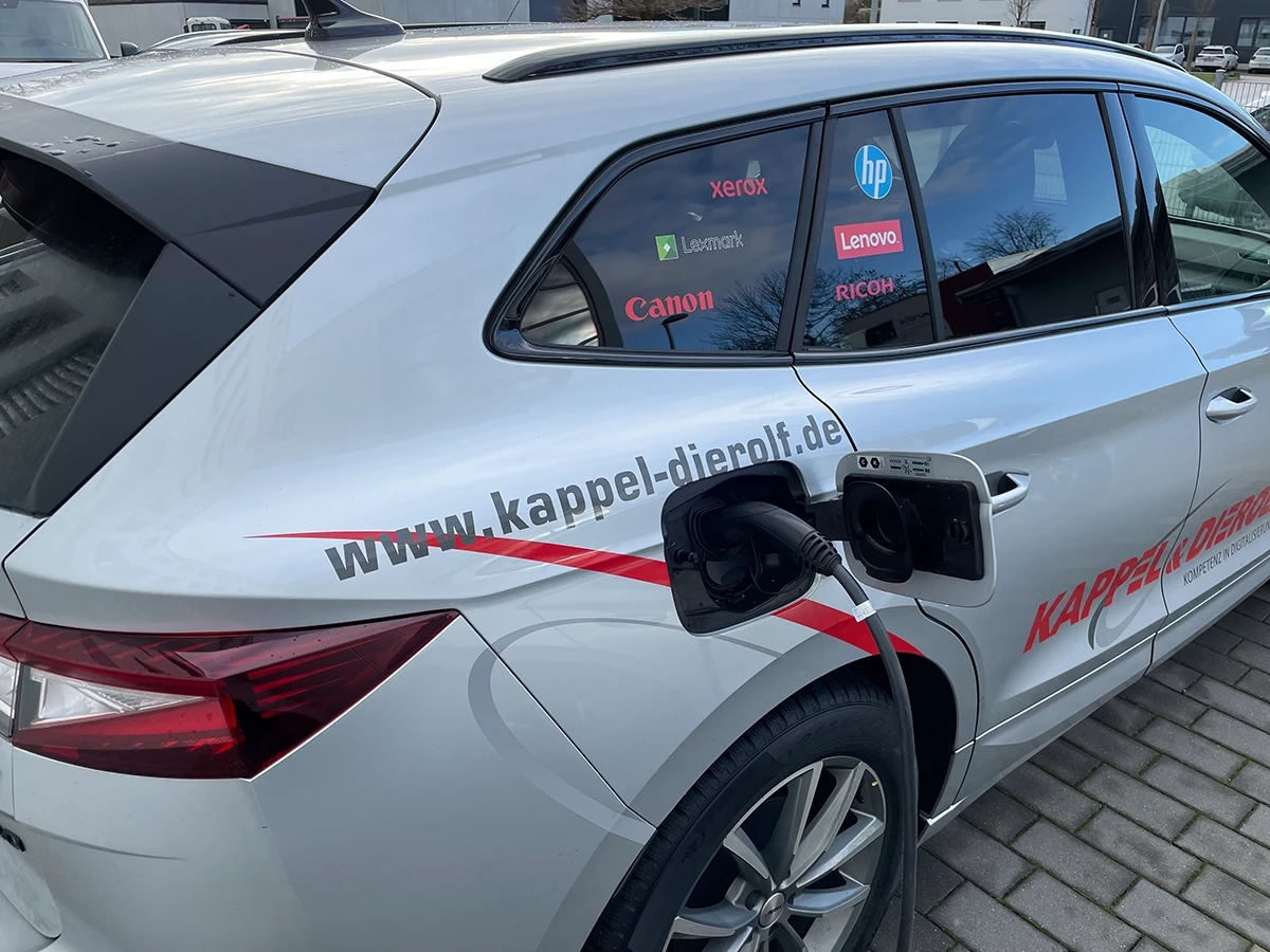 Silbernes Auto mit Ladekabel, Firmenlogo Kappel & Dierolf und Markenaufklebern, auf einem gepflasterten Parkplatz.