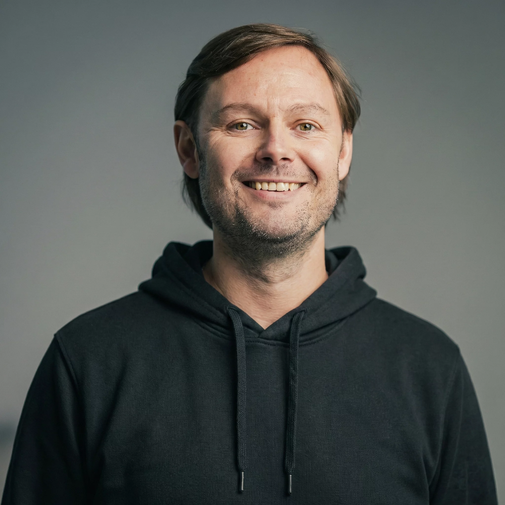 Eine Person lächelt in einem schwarzen Hoodie vor einem neutralen Hintergrund.