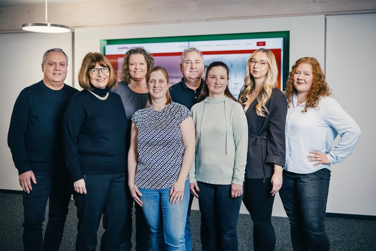 Gruppennaufnahme von acht Personen im Büro, lächelnd, vor einem Bildschirm mit Firmenwebsite im Hintergrund.