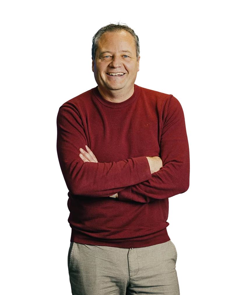 Ein lächelnder Mann in rotem Pullover steht mit verschränkten Armen vor weißem Hintergrund.