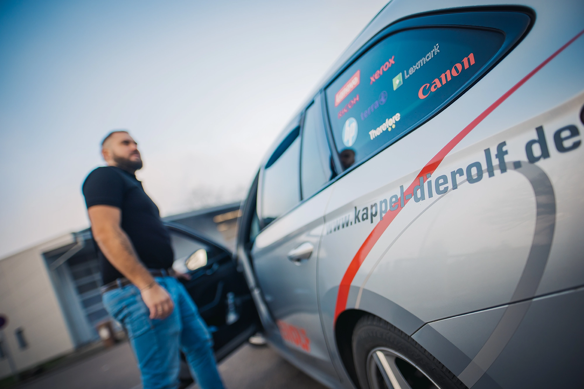Ein Mann steht neben einem grauen Auto mit Werbung und geöffneter Tür; im Hintergrund ein Industriegebäude.