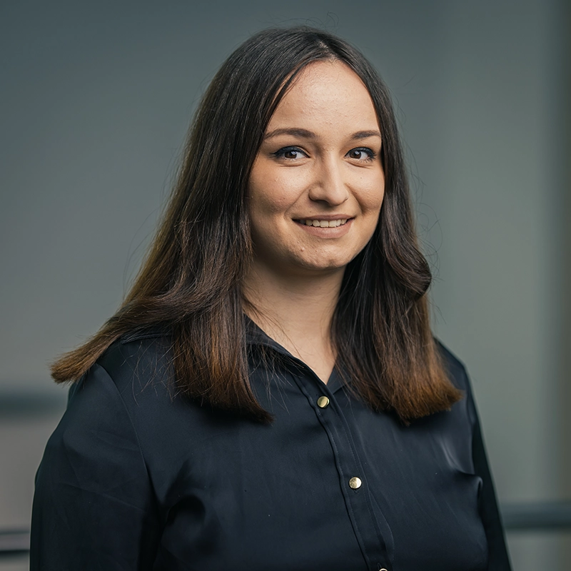 Eine Frau mit glattem, schulterlangem Haar trägt eine dunkle Bluse und lächelt in die Kamera. Hintergrund unscharf.