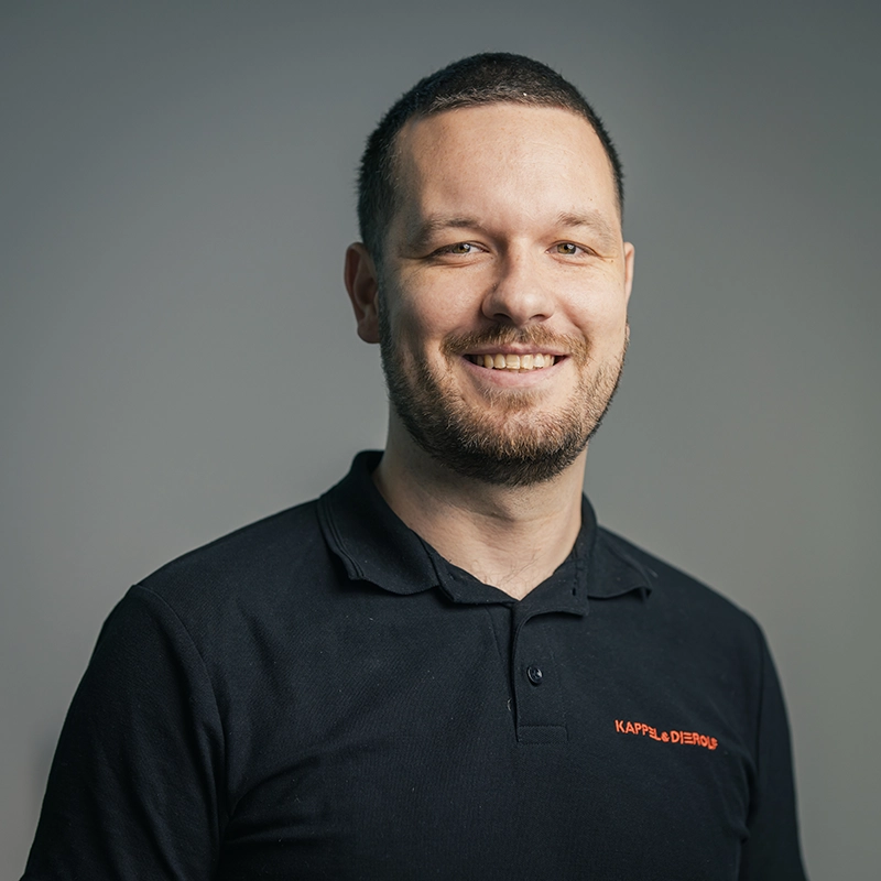 Ein Mann in dunkelblauem Poloshirt mit Kappel & Dierolf-Logo lächelt vor grauem Hintergrund.