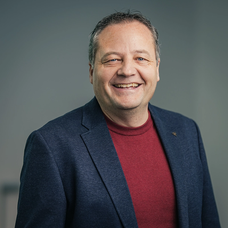Kappel_Potrait_Binder Ein lächelnder Mann in rotem Pullover und dunkelblauem Sakko vor grauem Hintergrund.