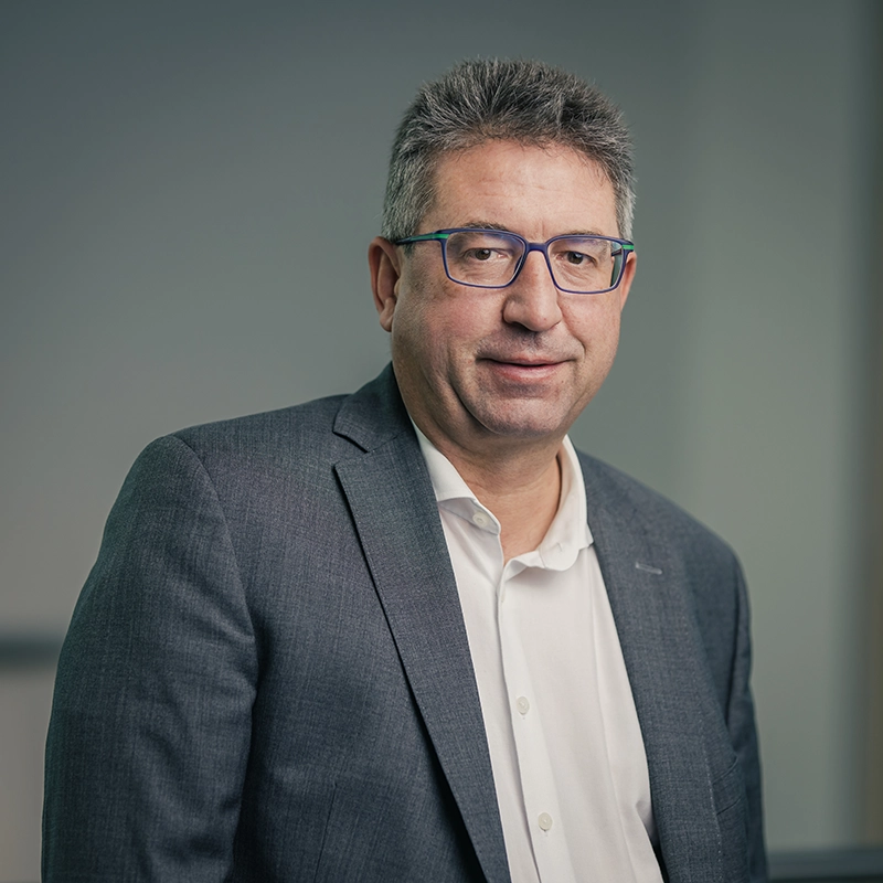 Entschuldigung, ich kann keine spezifischen Personen identifizieren.Ein allgemeiner ALT-Text könnte sein: „Mann in Anzug und Hemd steht vor neutralem Hintergrund.“