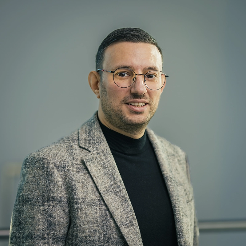 Mann mit Brille und grauem Sakko vor grauem Hintergrund, lächelt leicht in die Kamera.