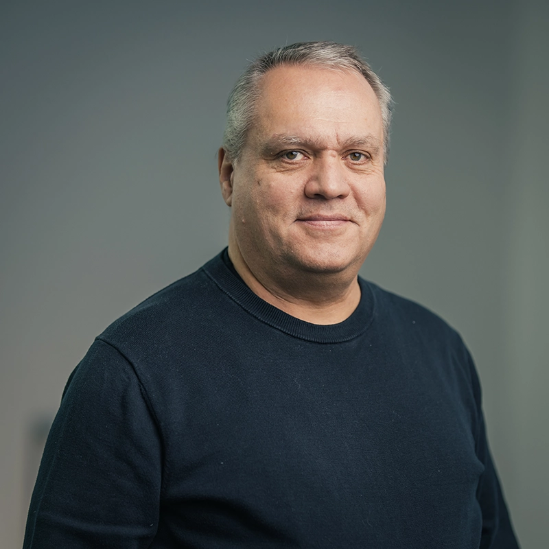 Eine Person in einem dunkelblauen Pullover lächelt leicht vor einem neutralen, grauen Hintergrund.