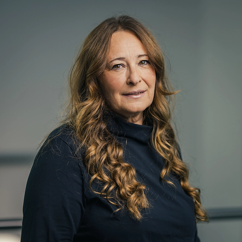 Eine Frau mit langen, lockigen Haaren blickt in die Kamera, trägt einen dunklen Pullover, vor neutralem Hintergrund.