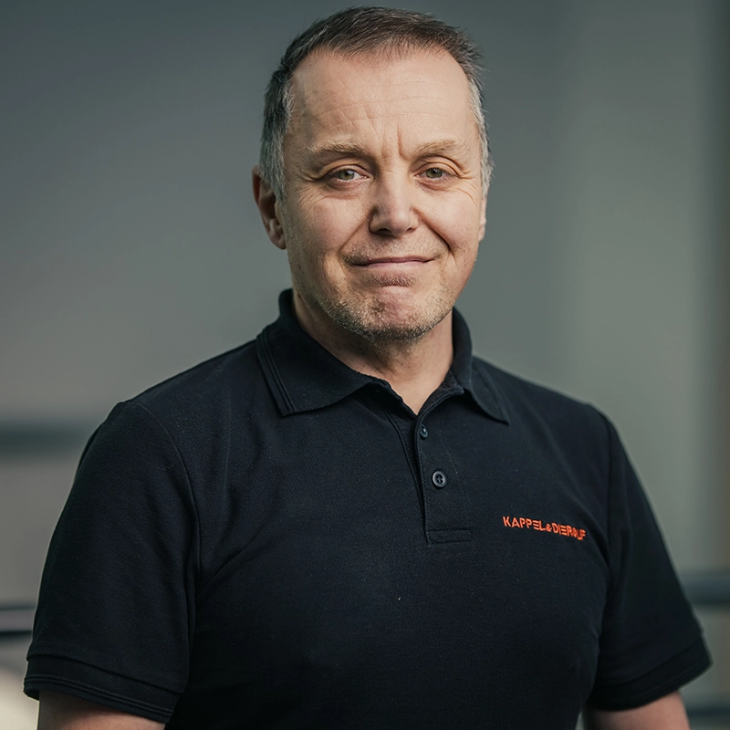 Ein Mann in einem schwarzen Polohemd vor einem neutralen, unscharfen Hintergrund lächelt leicht in die Kamera.