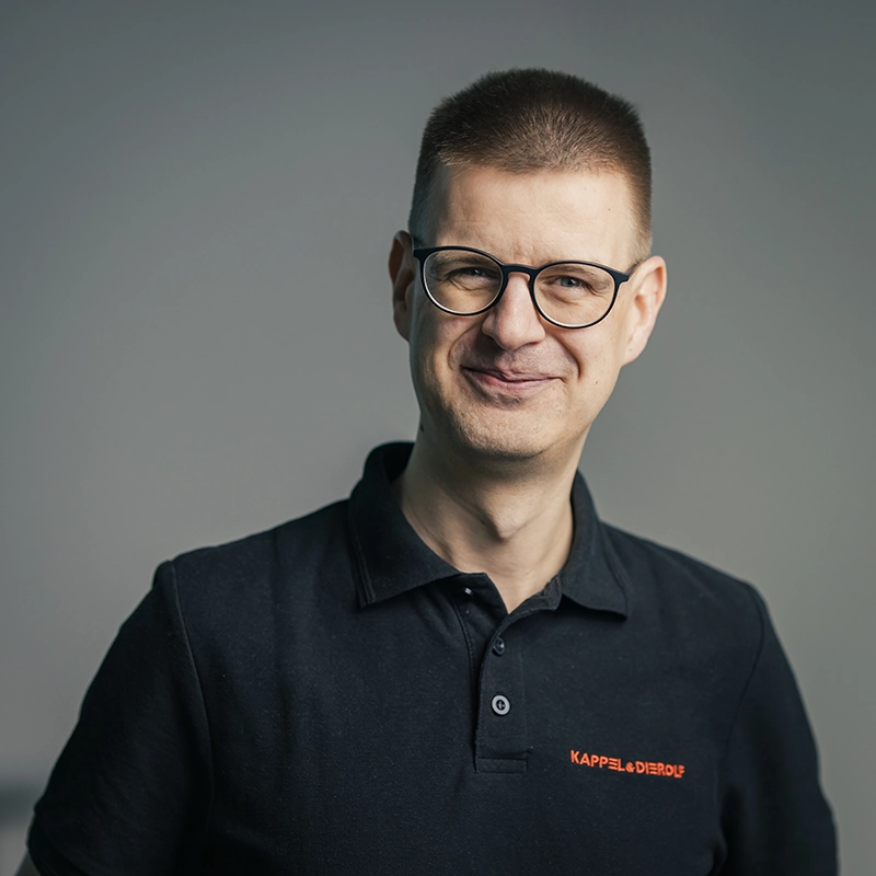 Person mit Brille und schwarzem Poloshirt lächelt vor grauem Hintergrund. Auf dem Shirt steht „KAPPELDIEROLF“.