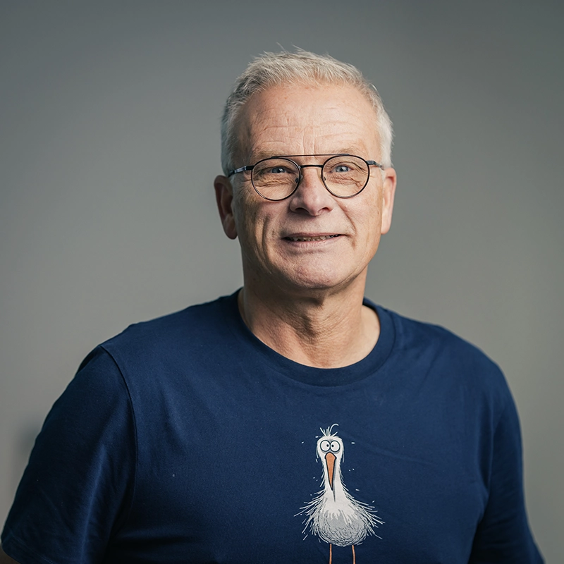 Ein älterer Mann mit Brille trägt ein dunkelblaues T-Shirt mit einem Cartoon-Vogel, lächelt vor neutralem grauem Hintergrund.