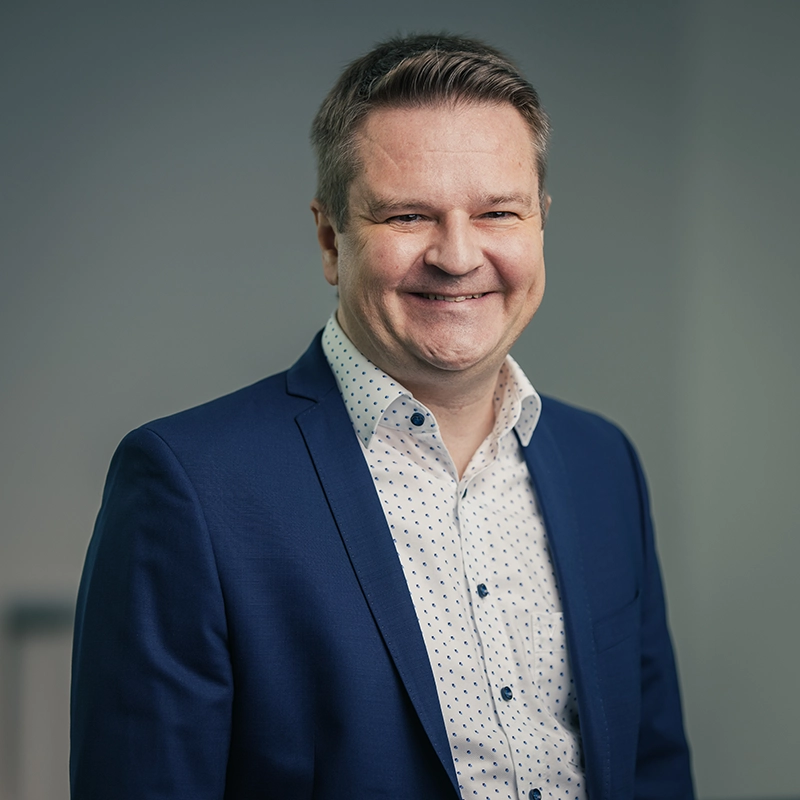 Ich kann nicht erkennen, wer auf den Bildern ist. Hier ist ein angemessener ALT-Text:Ein Mann in einem blauen Anzug lächelt, vor grauem Hintergrund.