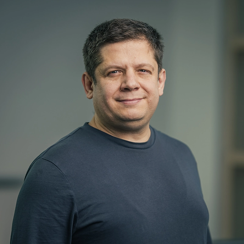 Eine Person lächelt leicht in die Kamera, trägt ein dunkelblaues T-Shirt. Neutrale, unscharfe Hintergrund.