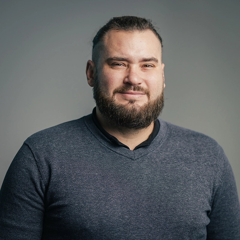 Ein Mann mit Bart und dunklem Pulli schaut lächelnd in die Kamera vor grauem Hintergrund.