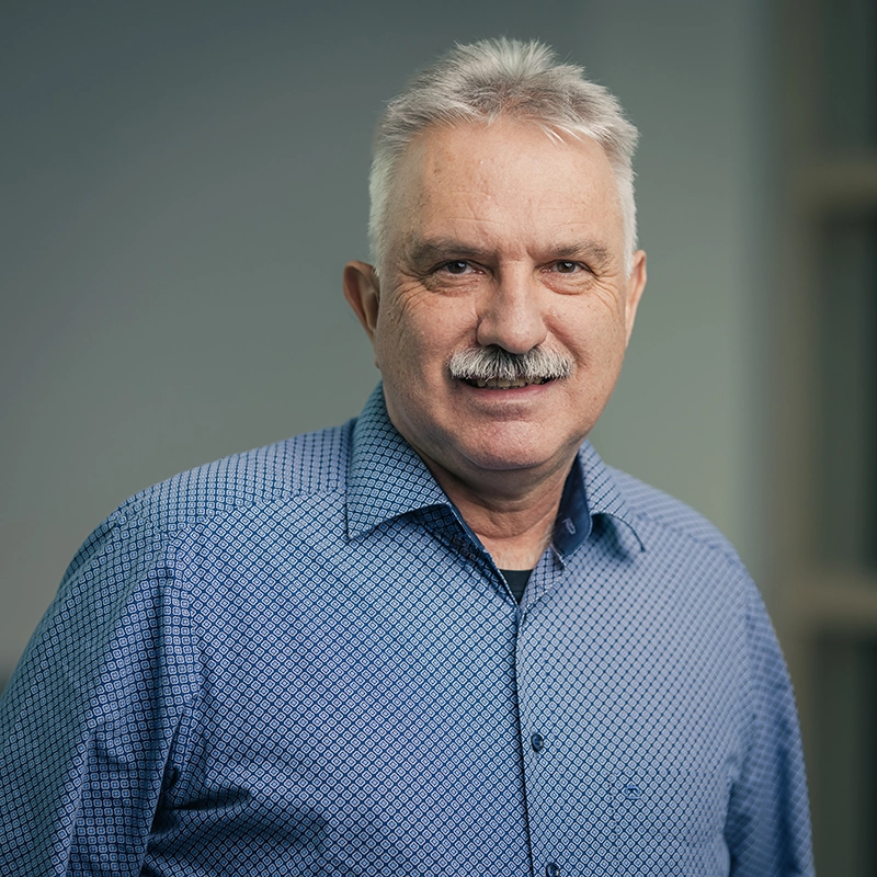 Älterer Mann mit grauen Haaren und Schnurrbart, trägt ein blau-gemustertes Hemd, lächelt freundlich in die Kamera.