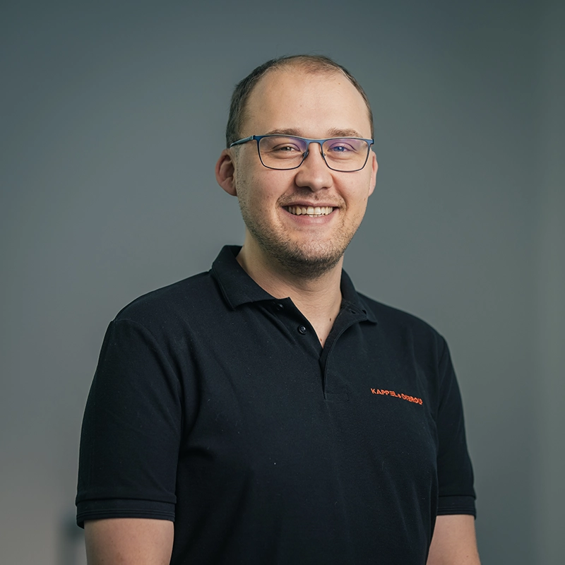 Eine lächelnde Person mit Brille in schwarzem Polohemd vor grauem Hintergrund.