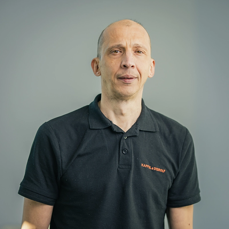 Eine Person im schwarzen Polohemd mit Firmenlogo steht vor einem neutralen Hintergrund.