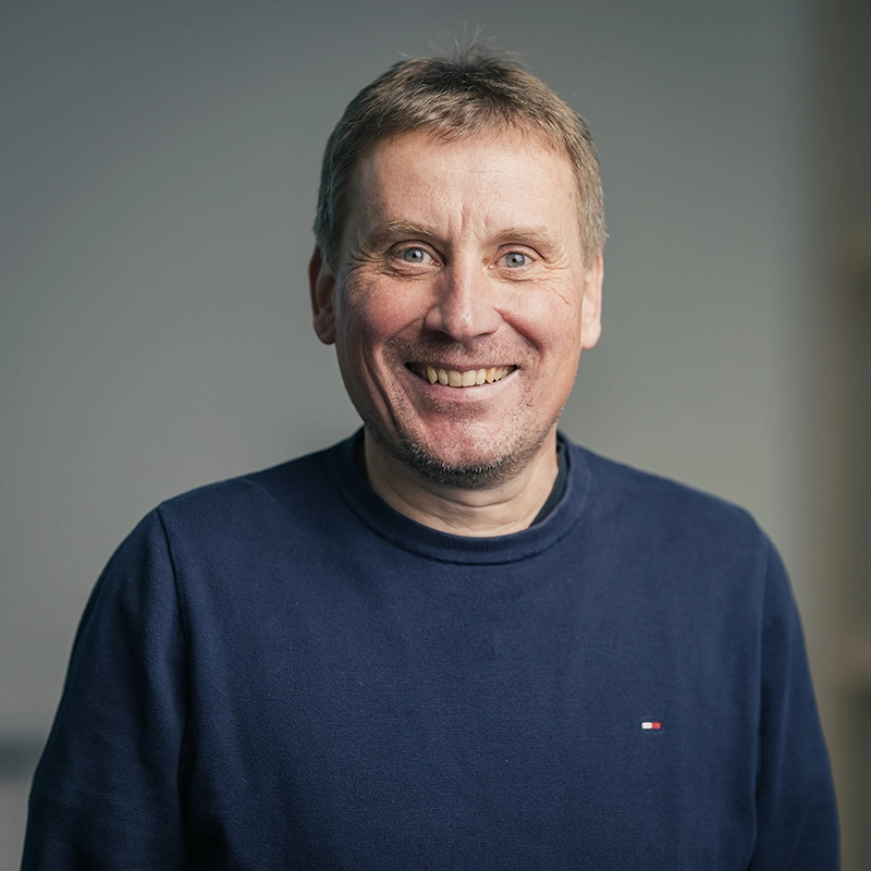 Ein lächelnder Mann in einem dunkelblauen Pullover vor neutralem Hintergrund, Blick in die Kamera.