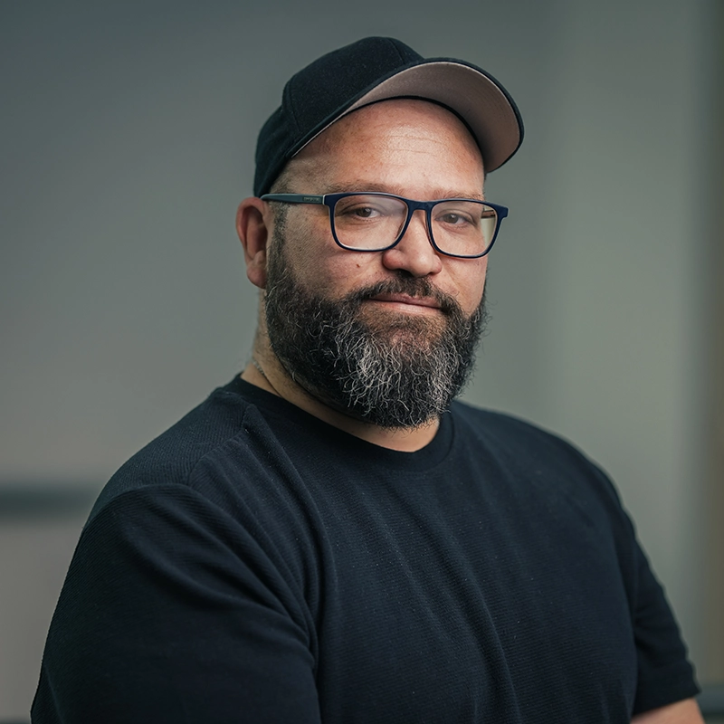 Ein Mann mit Bart und Brille trägt eine schwarze Mütze und schwarzes T-Shirt, schaut freundlich in die Kamera.