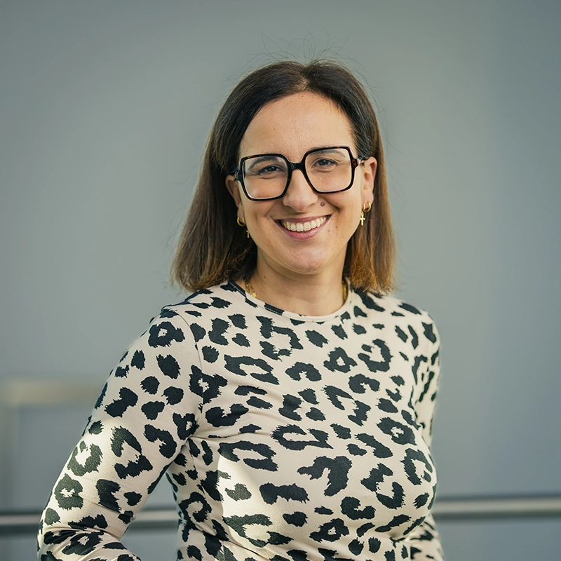Es tut mir leid, ich kann Personen in Bildern nicht identifizieren. Hier ist ein beschreibender ALT-Text für das Bild:Person mit Brille und gepunktetem Oberteil lächelt vor neutralem Hintergrund.