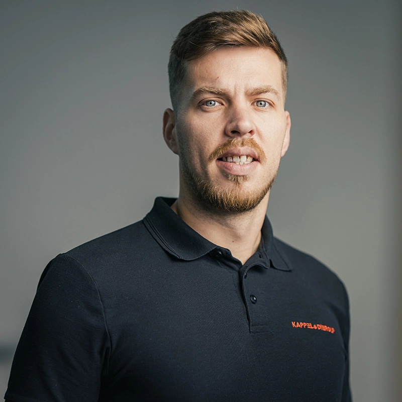 Eine Person mit Bart im schwarzen Polohemd steht vor grauem Hintergrund und blickt in die Kamera.