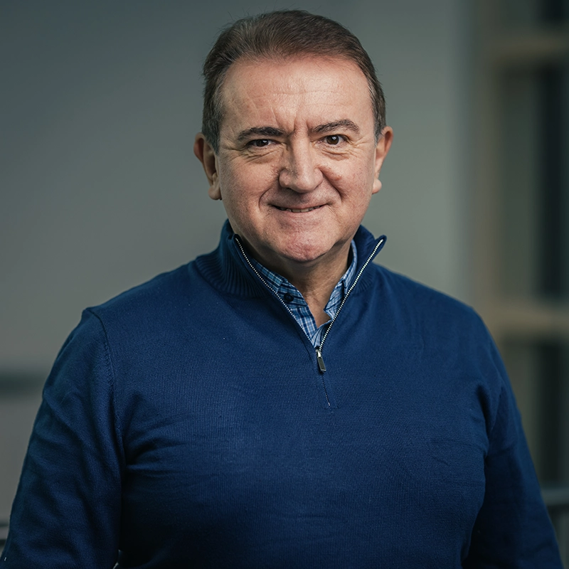 Ein lächelnder Mann in einem blauen Pullover und kariertem Hemd steht vor einem unscharfen Hintergrund.