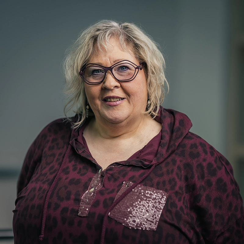 Eine lächelnde Frau mit blonden Haaren und Brille, trägt einen dunklen Pullover vor grauem Hintergrund.