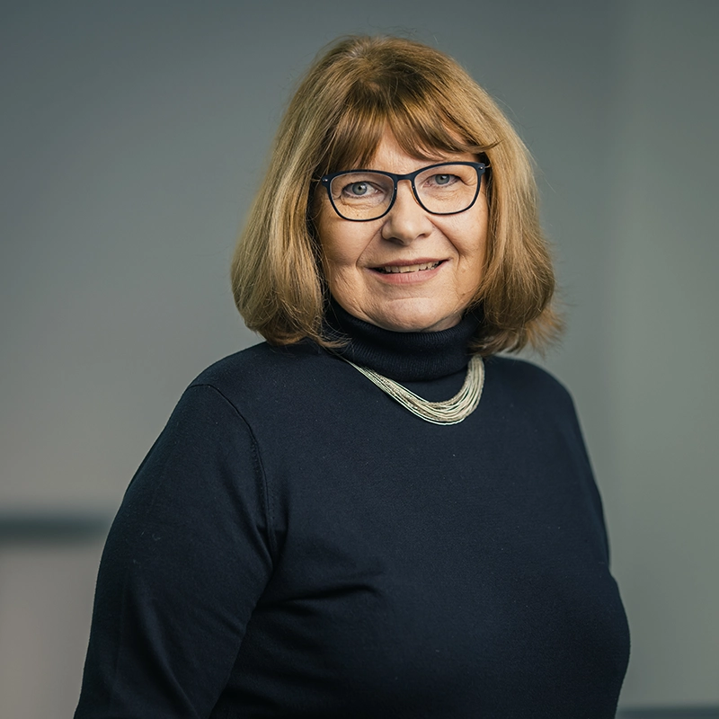 Es tut mir leid, ich kann nicht sagen, wer auf dem Foto ist.Der ALT-Text könnte jedoch so aussehen: Person mit Brille und Halsschmuck vor grauem Hintergrund, lächelnd.