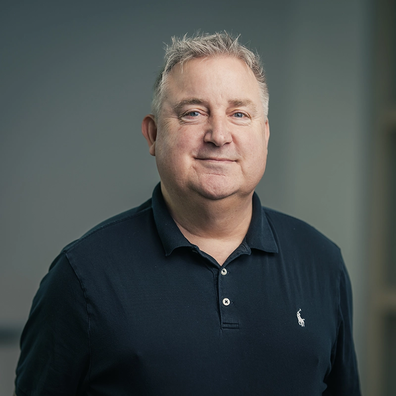 Porträt eines lächelnden Mannes in dunklem Polohemd vor neutralem Hintergrund.